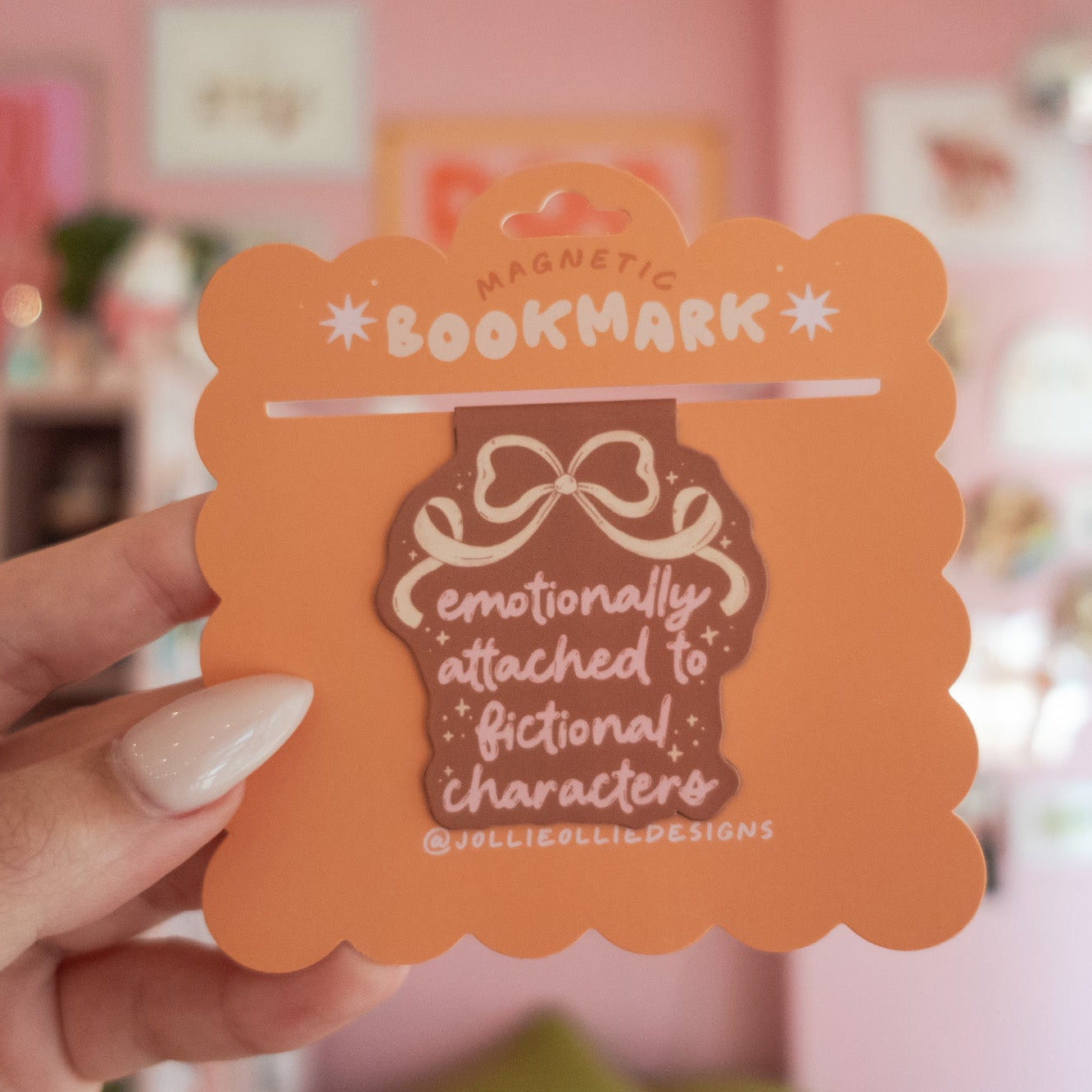 Brown magnetic bookmark with text that reads "emotionally attached to fictional characters" held by a hand against a blurred indoor background