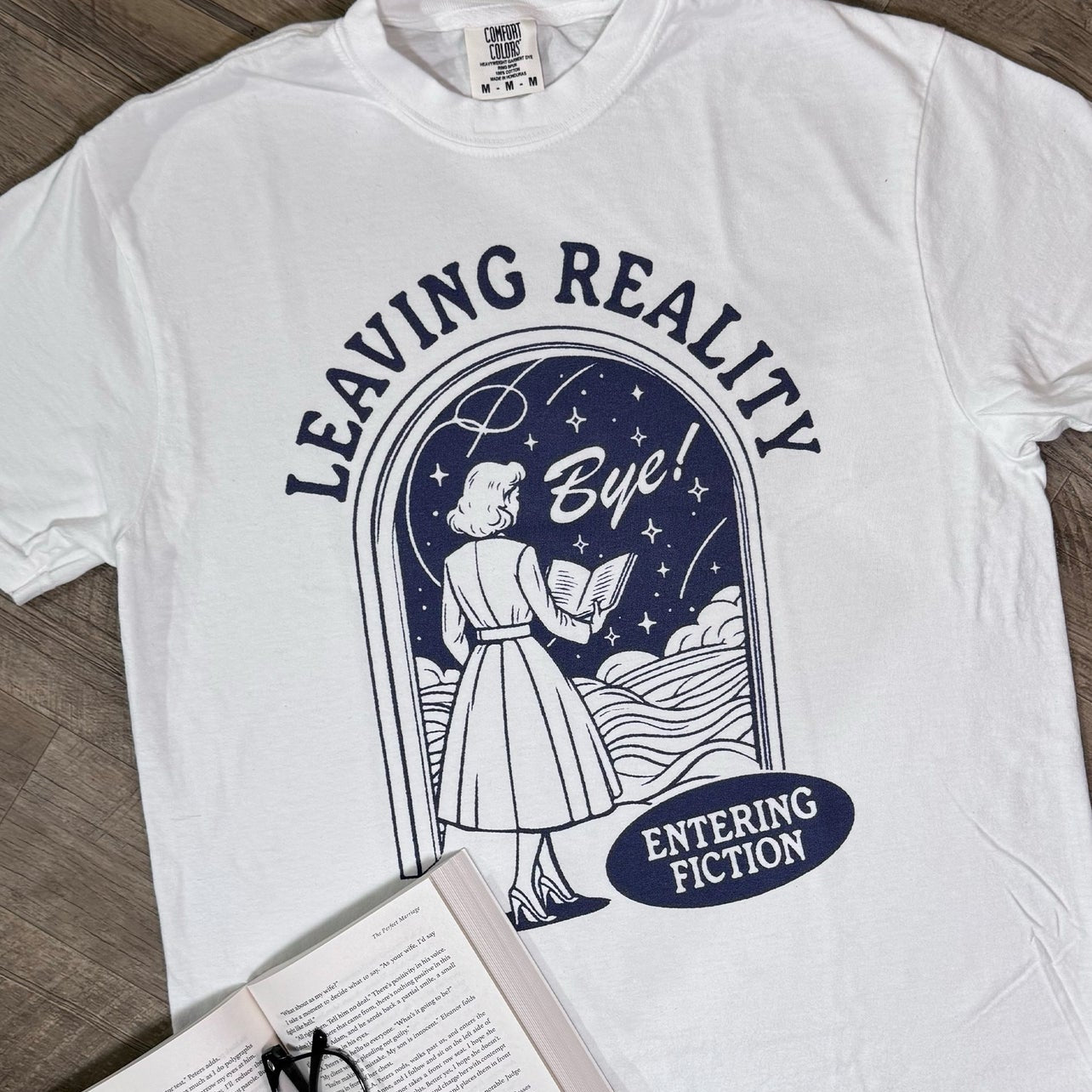 White t-shirt with graphic design of a woman and text 'Leaving Reality Bye! Entering Fiction' on a wooden surface with an open book and glasses.