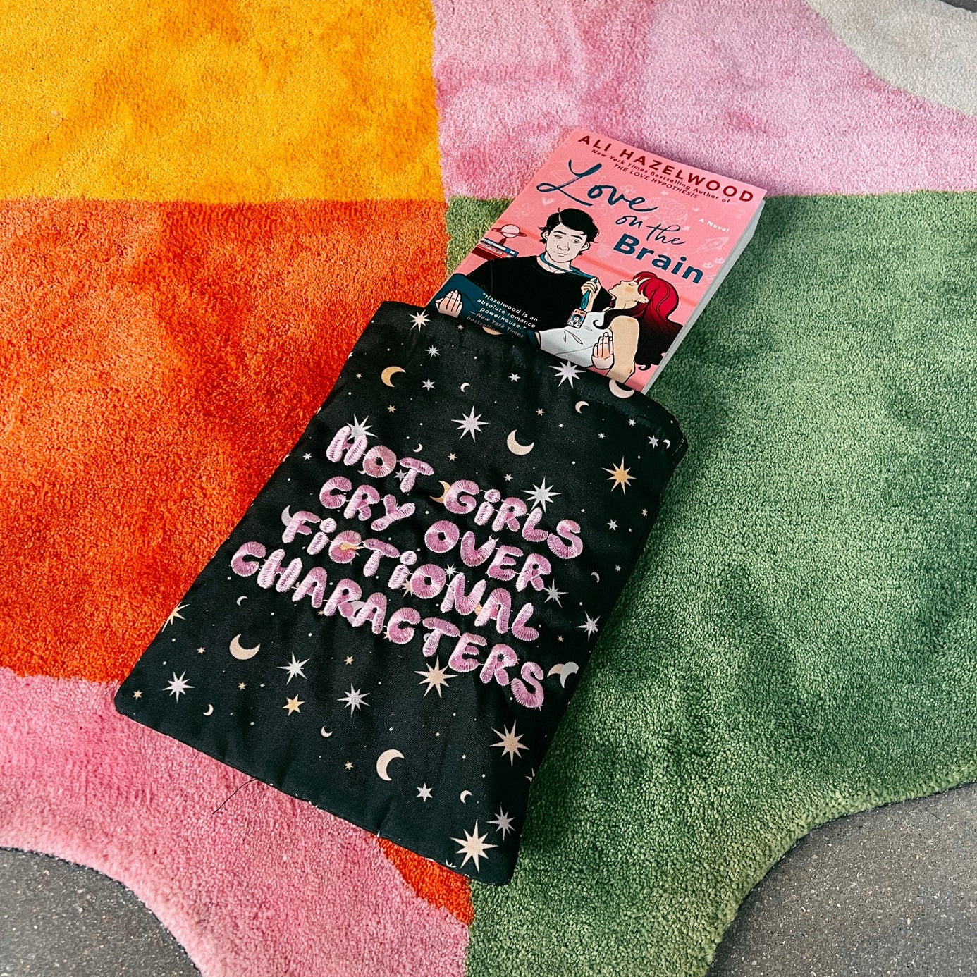 A book sliding out of a book sleeve on a colorful star-shaped rug. Book sleeve reads "hot girls cry over fictional characters"