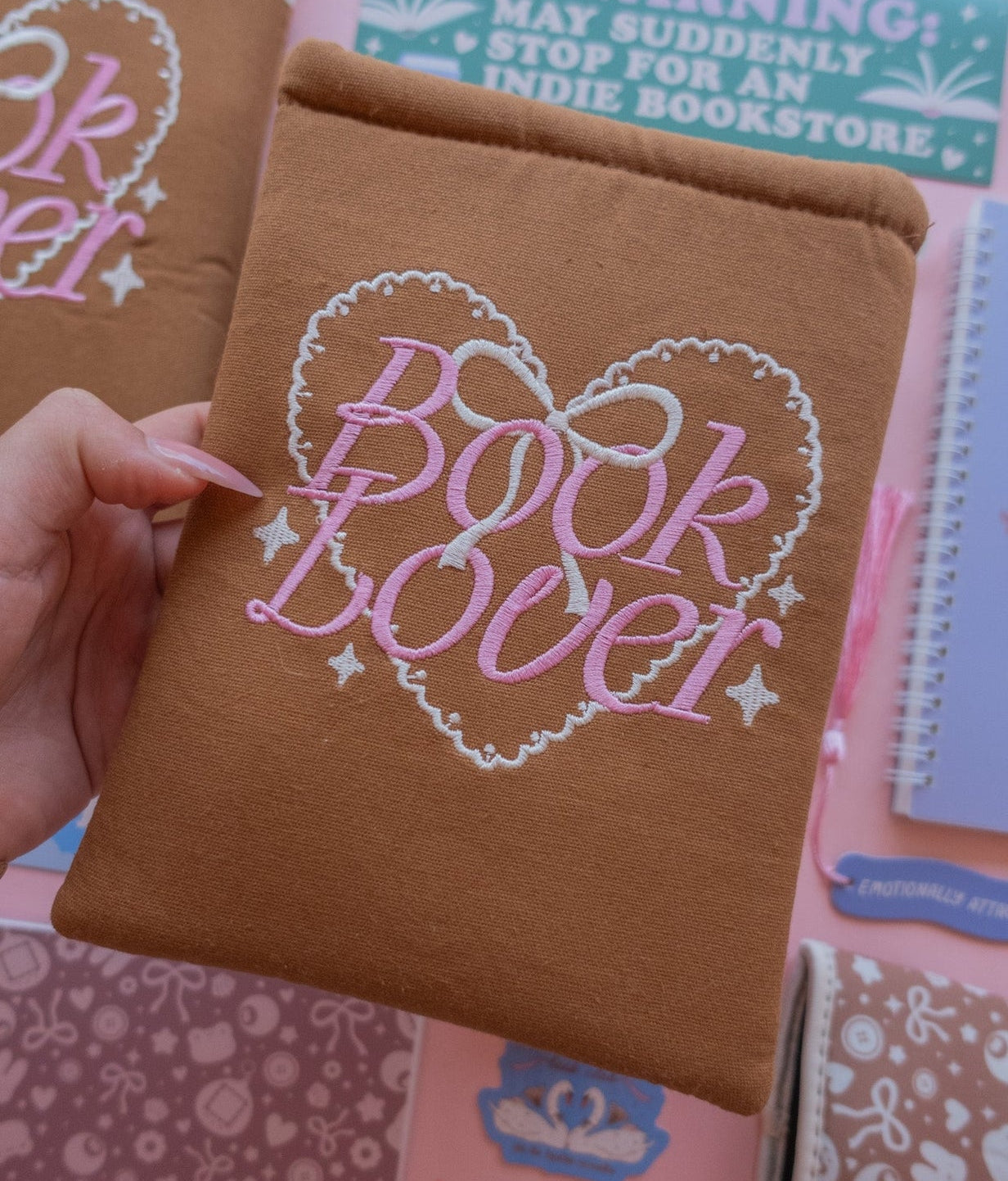 Brown book sleeve with 'Book Lover' embroidered text held by a hand, with other notebooks and stationery items in the background.
