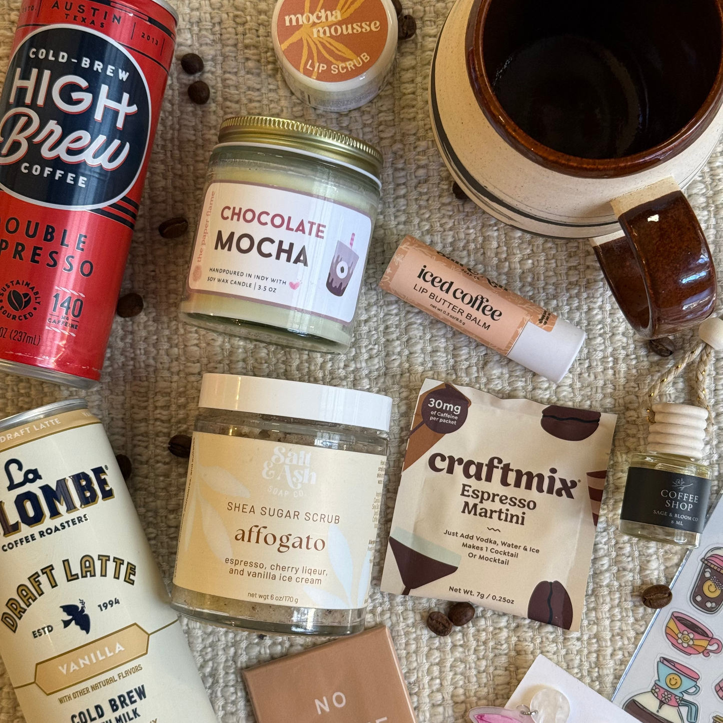 Collection of coffee-related products including a can of cold brew, a handmade coffee mug, and various themed items on a textured surface.