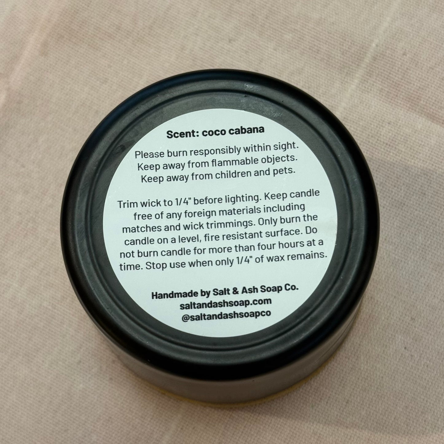 Candle lid with 'coco cabana' scent information on a beige fabric background