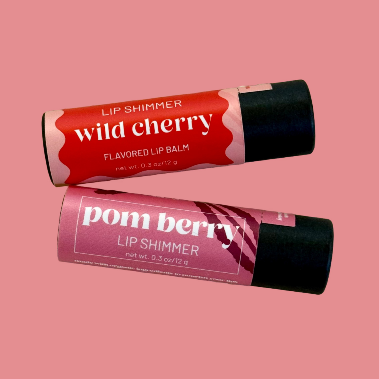 Two lip balm tubes, one labeled 'wild cherry' and the other 'pom berry', on a textured surface.