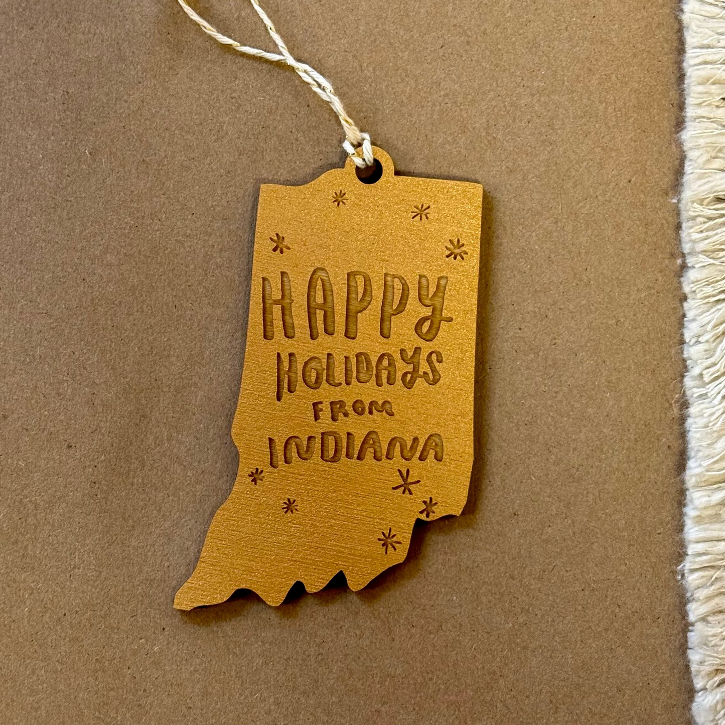 Wood ornament shaped like the state of Indiana with 'Happy Holidays from Indiana' text engraving - on a brown background.