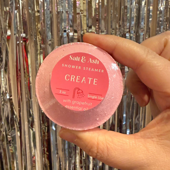Hand holding a pink shower steamer labeled 'Create' against a metallic tinsel background
